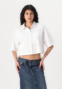 Camisa branca de botão cortada, com mangas curtas, colarinho e um bolso no peito, combinada com uma saia de ganga azul com uma abertura na frente.