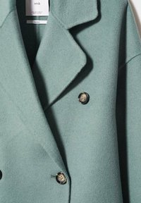 Manteau en laine à double boutonnage de couleur verte atténuée, avec un col cranté et deux boutons noirs à finition brillante. Tissu texturé.