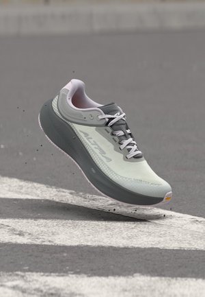 Zapatilla de correr gris claro y blanca con suela gruesa gris oscuro y cordones, suspendida sobre una carretera asfaltada con líneas blancas pintadas.
