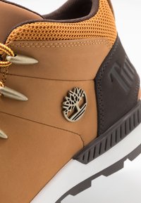 Scarpa da trekking in pelle marrone con dettagli in rete, dotata di logo metallico, cuciture decorative e suola in gomma testurizzata.