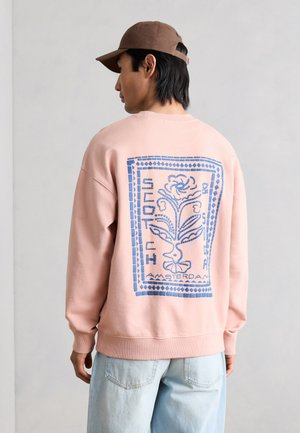 Homme portant un sweat-shirt rose avec un motif bleu floral et d'oiseau ainsi que les textes "SOUTH" et "AMSTERDAM" dans le dos, associé à un jean clair et une casquette marron.
