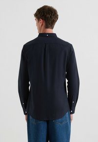 Camicia a maniche lunghe blu navy con colletto a punta e bottoni bianchi, caratterizzata da una texture liscia e un orlo arrotondato. Indossata con pantaloni in denim blu.
