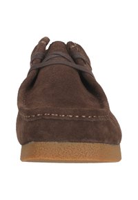 Ruskeakoonen mokkasiinikenkä, jossa on brodeerattua yksityiskohtaa ja nahkanyöriä. Litteä kumipohja, jossa on laikullinen pito. Etuosa korostaa avointa muotoilua.