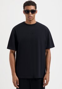 Zwarte t-shirt van zacht materiaal, met een ronde halslijn, korte mouwen en een subtiel reliëflogo aan de voorkant in het midden.