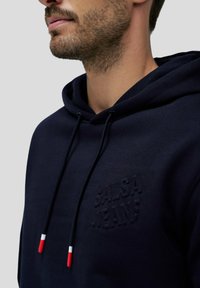 Felpa blu marina con una texture morbida, caratterizzata da un logo testurizzato e lacci con punte rosse, bianche e blu. Dettaglio della zona del collo.