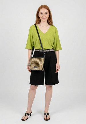 Frau trägt ein grünes V-Ausschnitt-Top, schwarze knielange Shorts, schwarze Sandalen mit Absatz und trägt eine beige gemusterte Crossbody-Tasche.