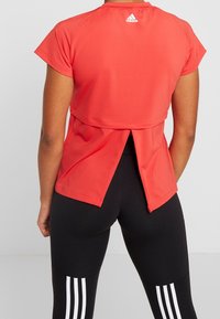 Červený sportovní top s krátkými rukávy a rozparkem na zadní straně. Vyrobený z hladkého, lehkého materiálu. Logo Adidas na krku.