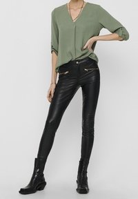 Top verde oliva a V con maniche arrotolate, abbinato a jeans skinny neri in pelle sintetica con dettagli in metallo dorato e stivaletti.
