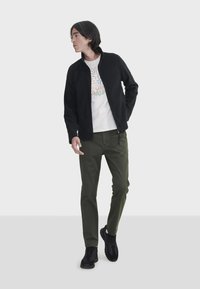 Schwarze Zip-Up-Jacke, weiße Grafik-T-Shirt, olivgrüne Hose und schwarze Sneaker. Glattes Material, lässige Passform, minimalistisches Design, ohne Muster.