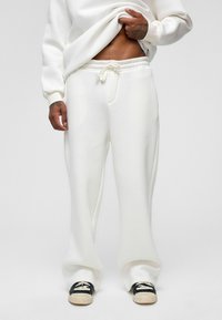 Witte, losse trainingsbroek met een elastische tailleband met trekkoord en zijzakken. Zachte textuur met een ontspannen silhouet, gecombineerd met zwarte schoenen.