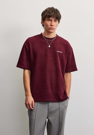 Giovane uomo che indossa una maglietta a maniche corte color granata con piccolo logo bianco, pantaloni grigi e due collane d'argento su uno sfondo chiaro e uniforme.