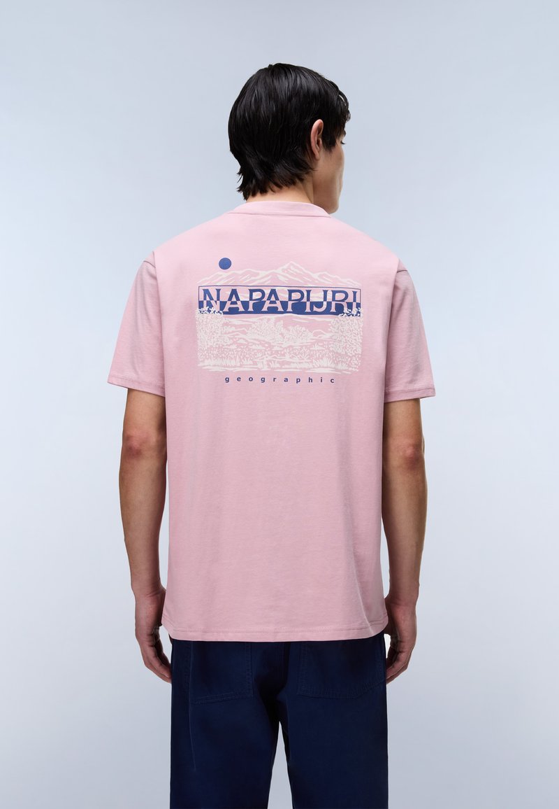 Homme de dos portant un T-shirt rose avec un graphique de montagne et le texte "NAPAPIJRI geographic" au dos, associé à un pantalon bleu foncé.