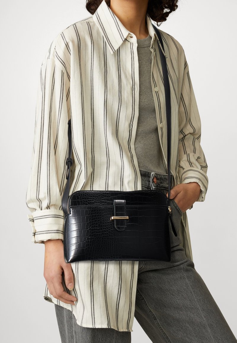 Frau in grauem Oberteil, gestreiftem beigen Hemd und Jeans, die eine schwarze, strukturierte Leder-Crossbody-Tasche mit goldenem Verschluss hält.