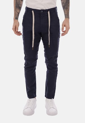 Hombre con pantalones ajustados azul marino con cordones blancos, combinados con zapatillas blancas y una camisa de color claro, de pie contra un fondo liso.