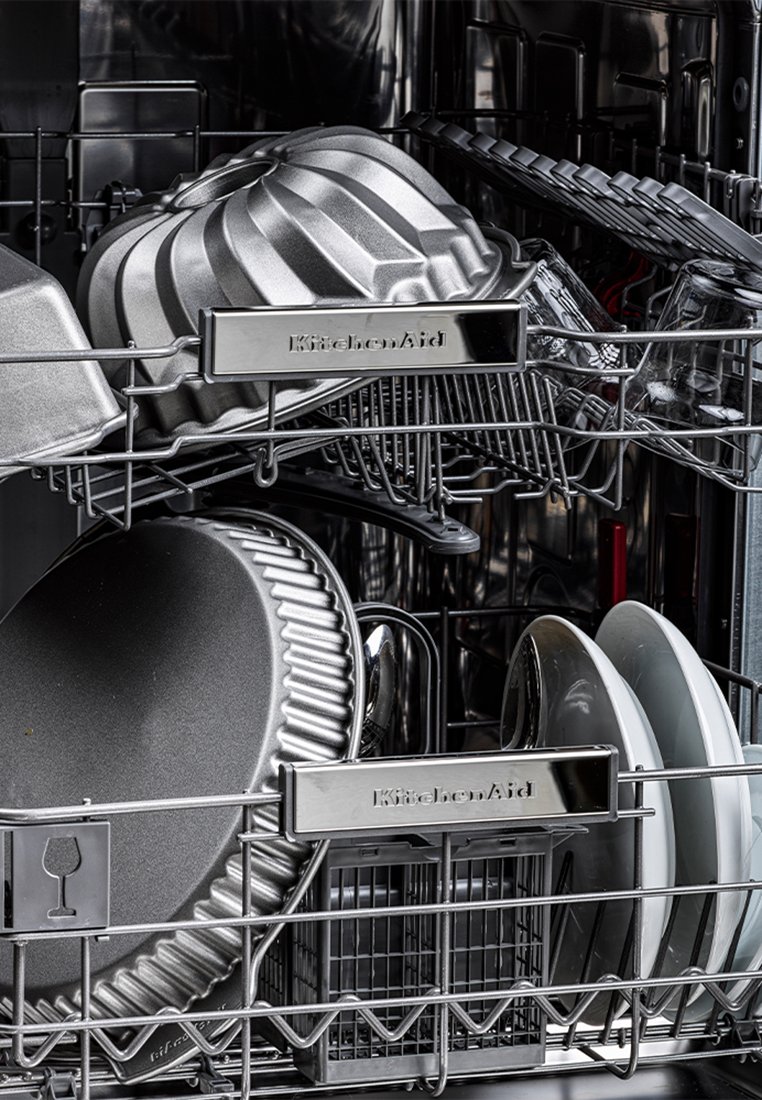 Zilveren metalen bakvormen en borden zijn gerangschikt in een roestvrijstalen vaatwasmachine rek met een KitchenAid merkdetail.