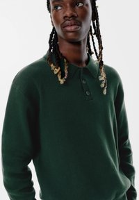 Tmavozelená pletená polo košeľa s golierom a tromi gombíkmi, s hladkou textúrou a voľným strihom, ktorú nosí osoba s dlhými vlasmi.