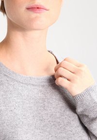 Maglione grigio a maglia con scollatura arrotondata e polsini a coste, caratterizzato da una consistenza morbida. Una mano che afferra delicatamente il colletto.