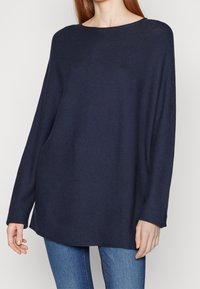 Pull ample bleu marine avec un col rond, manches longues, texture côtelée et ourlet droit, associé à un jean bleu.