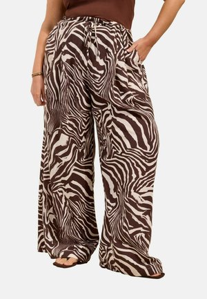 Bukser med vidde ben med brun og hvid zebra-print, kombineret med mørkebrunt top, en hånd i lommen og åbne sandaler synlige.