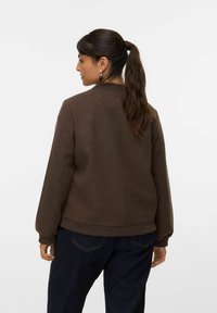 Sweatshirt marron avec un col montant, des manches longues et des poignets côtelés. Fabriqué en tissu texturé, associé à un jean bleu foncé.