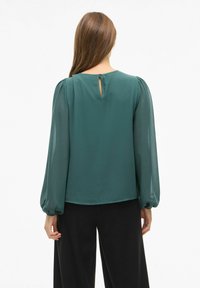 Blusa verde azulado con mangas largas y abullonadas, tela texturizada y un corte en forma de llave en la parte posterior. Combinada con pantalones negros de ajuste holgado.