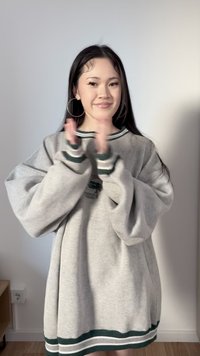 Νέα γυναίκα με μακριά σκούρα μαλλιά, φοράει φαρδιά γκρι μπλούζα με πράσινες ρίγες, χειροκροτεί και χαμογελά, στέκεται μέσα στον χώρο μπροστά σε έναν απλό τοίχο.