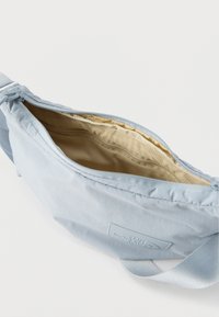 Ανοιχτό μπλε τσαντάκι ώμου με φερμουάρ ανοιχτό, μπεζ εσωτερική επένδυση και ρυθμιζόμενο λουράκι σε λευκό φόντο.