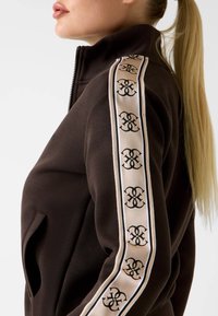 Brązowy rozpinany sweter z kieszeniami bocznymi, z kremowym paskiem z powtarzającymi się czarnymi wzorami logo wzdłuż rękawów. Miękka faktura.