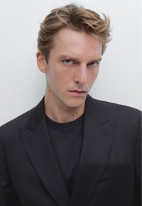 Blazer nero sopra una maglietta nera, con spalle strutturate e tessuto liscio. Capelli ondulati di media lunghezza, pelle chiara e tratti facciali marcati.