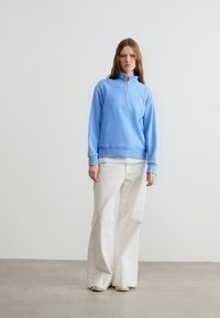 Γυναίκα που φοράει ανοιχτό μπλε φούτερ με φερμουάρ στο μισό μήκος, λευκό παντελόνι φαρδύ στο κάτω μέρος και λευκά παπούτσια, στέκεται μπροστά σε έναν απλό λευκό τοίχο.