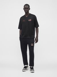 Άνδρας που στέκεται φορώντας μαύρο oversized κοντομάνικο πουκάμισο και μαύρο παντελόνι με λευκά και μαύρα αθλητικά παπούτσια σε απλό λευκό φόντο.