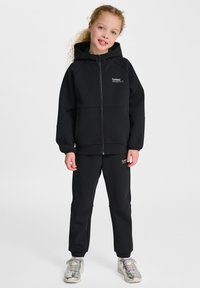 Svart zip-up hoodie med huva och framfickor, kombinerad med matchande svarta joggers. Båda har minimal logotypdetalj och en mjuk textur.