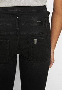 Jeans negros ajustados con una textura suave, que cuentan con bolsillos traseros y una etiqueta negra de marca en la cintura con un detalle metálico.