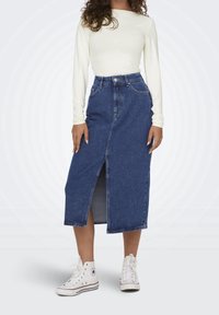 Denim midi-kjol med framfickor, hög midja och blå färg. Matchas med en långärmad vit topp och vita högklackade sneakers.