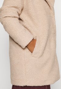 Beige fleecejacka med en texturerad, fluffig yta, med en sidficka och avslappnad passform. Synliga vikta ärmar.