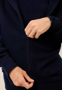 Laivasto sininen vetoketjullinen neule, ribbitettua tekstiiliä, etuosa vetoketjulla ja tiukat hihansuut. Kädet pitävät vetoketjusta ja kankaan helmasta.