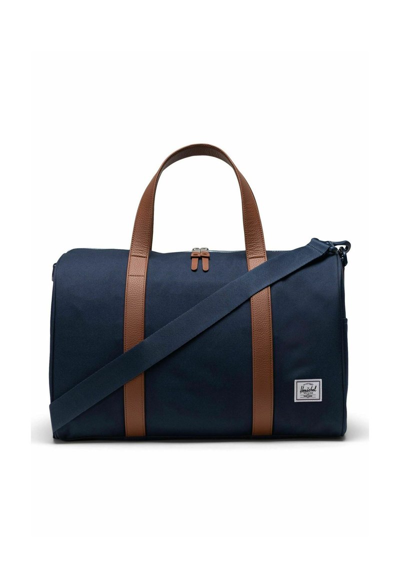 Sac de sport en toile bleu marine avec des poignées et des accents en cuir marron, fermeture éclair et bandoulière réglable. Comprend un petit patch logo à l'avant.