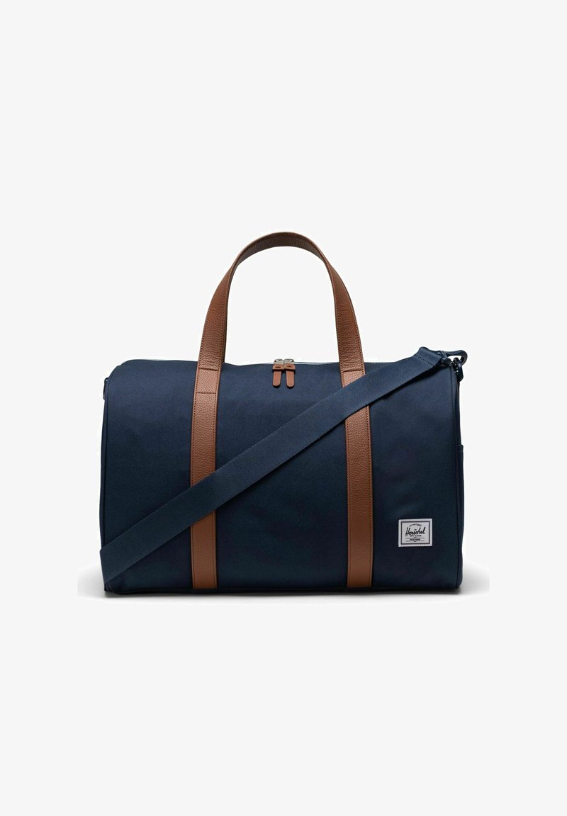 Sac de sport en toile bleu marine avec des poignées et des accents en cuir marron, fermeture éclair et bandoulière réglable. Comprend un petit patch logo à l'avant.