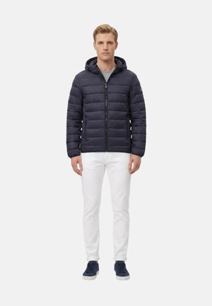 Giovane uomo in piedi che indossa una giacca trapuntata con cappuccio blu navy, pantaloni bianchi, camicia grigia e sneakers blu navy su sfondo bianco.