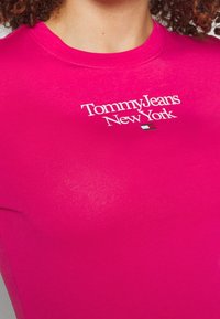 Detailní záběr na osobu v jasně růžovém tričku s bílým textem "Tommy Jeans New York" a malou vlajkou červené, bílé a modré barvy.