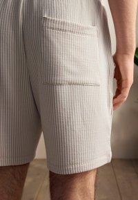 Light beige shorts with vertical stripes, featuring a side pocket and a textured fabric. Length extends to mid-thigh. Wooden floor background.
