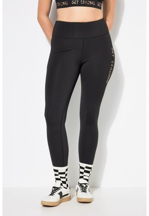 Leggings neri con vita alta riportano la scritta "GET STRONG" in oro sulla gamba sinistra. Abbinati a calzini a scacchi bianchi e neri e sneakers bianche.