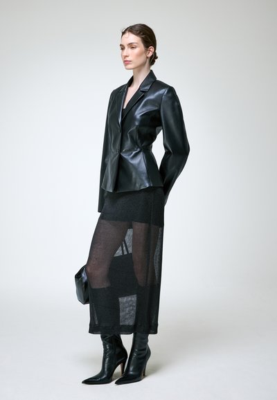 Femme portant un blazer en cuir noir, une jupe noire transparente sur un short, des bottes à talons noires, et tenant un petit sac à main noir sur un fond uni.