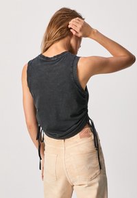 Svart ärmlös topp med rundad halsringning, samlade sidor och knytningar. I kombination med ljus beige jeans som visar en diskret fickdesign.