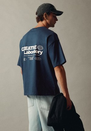 Homme portant une casquette marine, un t-shirt marine avec un texte blanc "Creative Laboratory", un jean bleu clair, tenant une veste noire sur un fond uni.