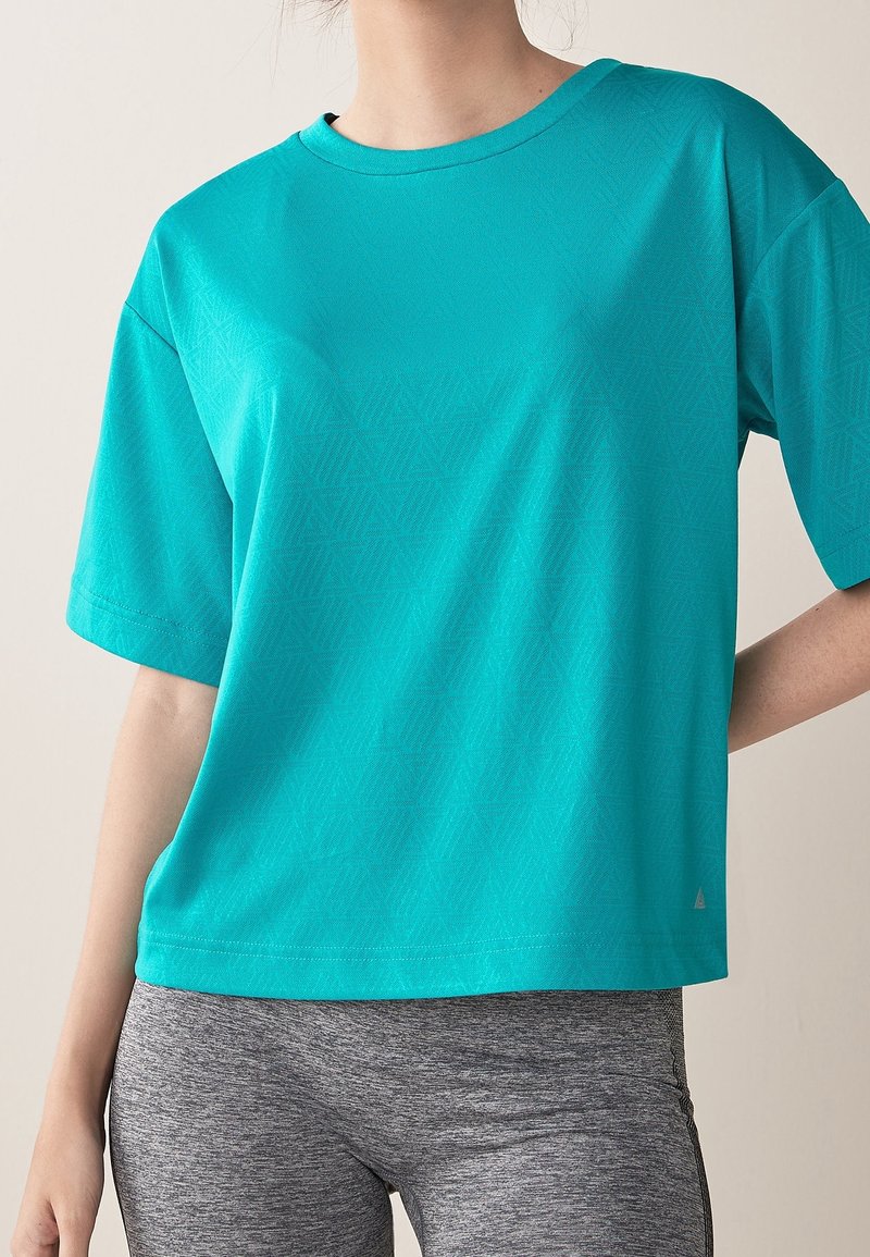 Frau trägt ein türkisfarbenes kurzärmliges Shirt mit dezenten geometrischen Mustern und graue enganliegende Leggings, steht mit einer Hand auf der Hüfte.