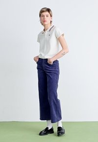 Chemise polo blanche avec col rayé, pantalon large en velours côtelé bleu marine, mocassins noirs, et chaussettes blanches, sur fond clair.