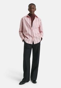 Frau steht mit den Händen in den Taschen. Sie trägt ein rosa gestreiftes Hemd über einem kastanienbraunen Rollkragenpullover, schwarze weit geschnittene Hosen und schwarze Schuhe.