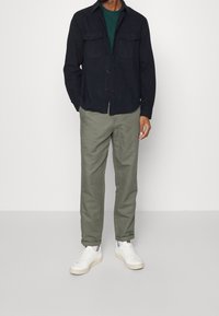 Uomo che indossa una giacca a camicia blu scuro, una maglietta verde, pantaloni verde oliva e sneakers bianche, in piedi contro uno sfondo bianco uniforme.