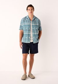 Kortærmet skjorte med et blåt og hvidt geometrisk mønster, kombineret med mørke shorts. Model iført afslappede sko, stående mod en lys baggrund.